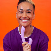 A person with short hair and tattoos is smiling while holding the Cute Little Fuckers Jix - Lavender, which features an ergonomic handle. They are wearing a purple shirt and earrings, standing against an orange background that accentuates the pleasurable vibe of the scene.